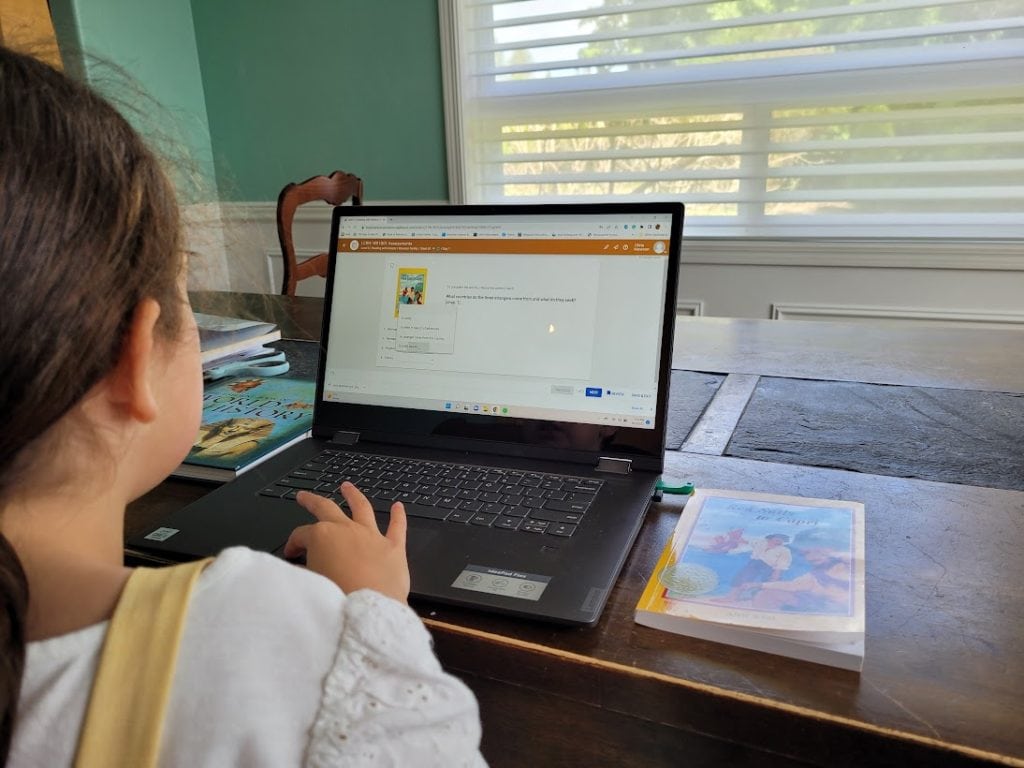Little girl answering questions on her laptop as part of BookShark Virtual. The book Red Sails to Capri is sitting next to the computer.