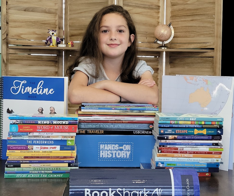 little girls standing behind BookShark Reading with History C curriculum stacked in piles